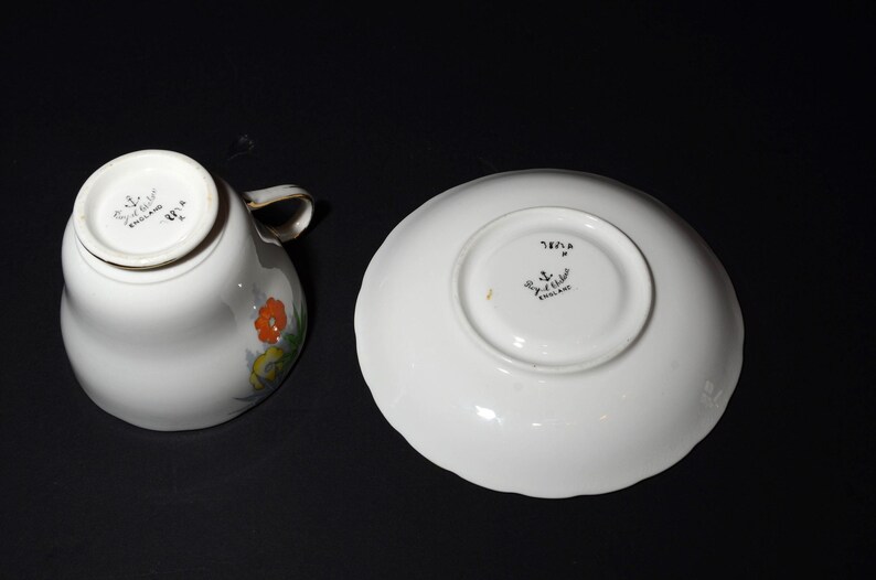 May include: White teacup and saucer set with floral design. The teacup has a handle and a floral pattern in orange, yellow, and green. The saucer is round with a scalloped edge. Both pieces have markings that say "England".