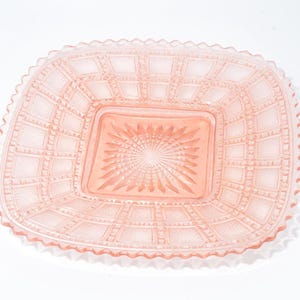 May include: A pink, rectangular glass dish with a textured grid pattern and a central square with a starburst design. The dish has a scalloped edge. A decorative piece for serving or display.