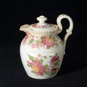 May include: Cream-colored ceramic creamer with a lid, adorned with pink, red, and green floral patterns. The creamer has a curved handle and a spout for pouring. The lid is topped with a small knob.