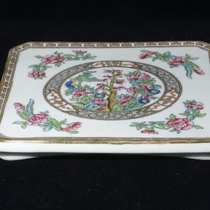 May include: A rectangular, white ceramic trinket box with a gold trim. The lid features a central oval design with a tree and floral motif in pink, blue, and green. Pink floral and bird accents are placed around the edges.