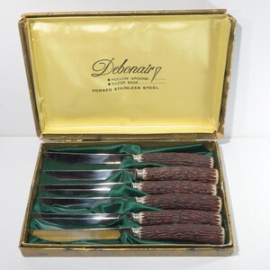 May include: A set of six stainless steel steak knives with brown, textured handles, presented in a vintage box. The box has a gold interior and the text "Debonair" is printed on the inside of the lid.