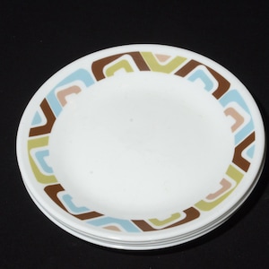 Puede incluir: Una pila de platos redondos blancos con un patrón decorativo en el borde de formas geométricas repetitivas en tonos marrón, azul claro, beige y verde oliva. Los platos tienen un acabado liso y brillante y un diseño sencillo en el borde.