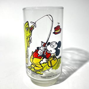 K&ouml;nnte beinhalten: Ein klares Glas mit einer Cartoon-Illustration von Mickey Mouse beim Angeln. Mickey tr&auml;gt eine rote Jacke und gelbe Hose, mit einem gelben Fisch an der Leine. Ein roter Hut und eine Angelkiste sind ebenfalls zu sehen. Das Glas ist mit "Walt Disney Productions" gekennzeichnet.