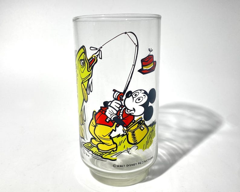 K&ouml;nnte beinhalten: Ein klares Glas mit einer Cartoon-Illustration von Mickey Mouse beim Angeln. Mickey tr&auml;gt eine rote Jacke und gelbe Hose, mit einem gelben Fisch an der Leine. Ein roter Hut und eine Angelkiste sind ebenfalls zu sehen. Das Glas ist mit "Walt Disney Productions" gekennzeichnet.