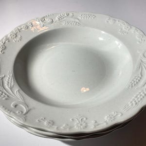 May include: A set of three white ceramic soup bowls with a raised floral design. The bowls have a scalloped edge and a deep well.
