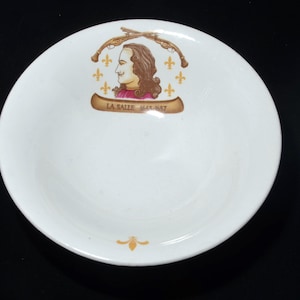 May include: A white ceramic bowl featuring a central graphic of a historical figure in profile, flanked by two crossed muskets and gold fleur-de-lis symbols. Below the portrait, a tan banner displays the text LA SALLE 1643-1687 in dark lettering.