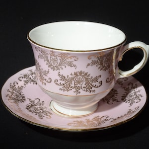 Op de afbeelding: Een roze theekopje en schotelset met een gouden bloemenpatroon. Het kopje heeft een witte binnenkant en een afgeronde handgreep.