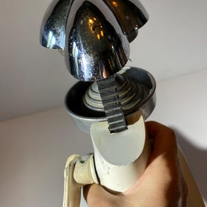 May include: A vintage white and chrome manual citrus juicer with a ribbed, black rubber belt. The juicer has a chrome top with a five-pointed star design.