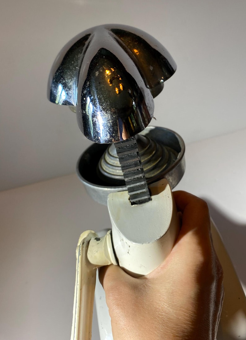May include: A vintage white and chrome manual citrus juicer with a ribbed, black rubber belt. The juicer has a chrome top with a five-pointed star design.