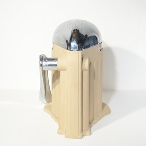 May include: A vintage manual citrus juicer with a chrome top and a beige base. The juicer has a handle on the side and a spout for pouring juice.