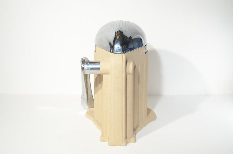 May include: A vintage manual citrus juicer with a chrome top and a beige base. The juicer has a handle on the side and a spout for pouring juice.