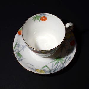 May include: White teacup and saucer set with a floral and bamboo design. The teacup and saucer have a gold trim. The teacup has a handle. The floral design includes orange, yellow, and green accents.