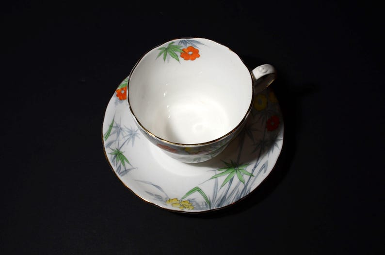 May include: White teacup and saucer set with a floral and bamboo design. The teacup and saucer have a gold trim. The teacup has a handle. The floral design includes orange, yellow, and green accents.