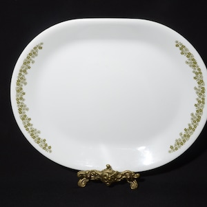 以下が含まれることがあります： 縁に緑色の花柄が施された白い楕円形のサービングプレート。プレートは金色の金属製のスタンドの上に置かれています。