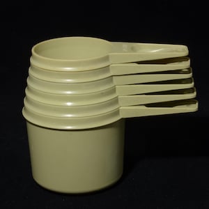 Peut inclure: Un ensemble de six tasses et cuillères à mesurer beiges, empilées. Les tasses sont cylindriques avec un large bord, et les cuillères s'étendent sur le côté. L'ensemble est en plastique de couleur claire.