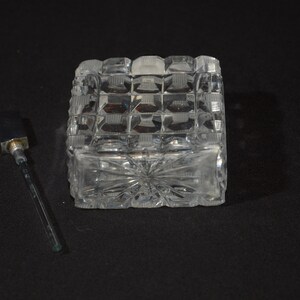 May include: A clear glass bottle with a square base and a decorative pattern. The bottle has a glass stopper with a metal top.