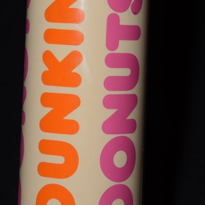 May include: A cylindrical container with the words "DUNKIN' DONUTS" printed vertically in orange and pink. The background is a light beige color, and the text is bold and easy to read. The container is against a black background.