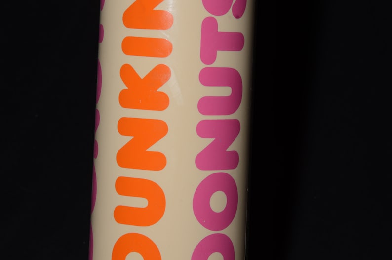 May include: A cylindrical container with the words "DUNKIN' DONUTS" printed vertically in orange and pink. The background is a light beige color, and the text is bold and easy to read. The container is against a black background.
