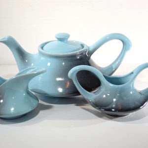 Puede incluir: Una tetera y dos jarras de crema de cerámica azul y negra. La tetera tiene un cuerpo redondeado y un pico. Las jarras tienen un asa curva y un pico.