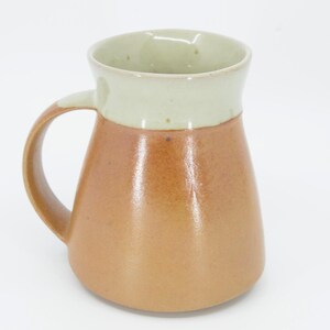 May include: A handmade ceramic mug with a rounded base and a large handle. The mug features a two-tone design, with a light beige rim and a warm brown body. The interior is a light green color.