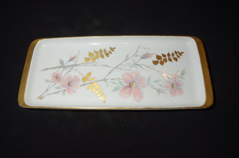 May include: Rectangular white ceramic tray with a gold rim. The tray is decorated with pink flowers, green leaves, and gold-colored leaf accents. A decorative piece for serving or display.
