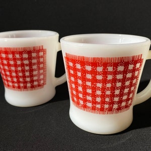 Może przedstawiać: Dwa białe ceramiczne kubki z czerwono-białym wzorem w kratkę. Kubki mają zaokrąglony kształt i uchwyt.