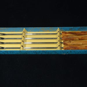 Può includere: Un set di sei forchette da fonduta in una scatola blu. Le forchette hanno denti e steli in acciaio inossidabile, con manici in legno. La scatola ha un interno giallo e un esterno blu con un motivo strutturato. I manici misurano circa 10 cm.