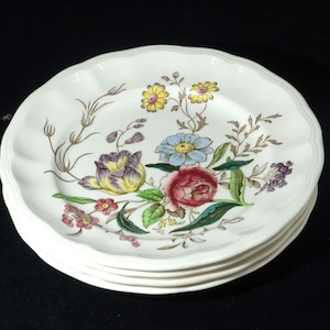 May include: Four white ceramic plates with a floral design. Each plate has a scalloped edge and is decorated with yellow, blue, purple, and red flowers, green leaves, and brown stems. The plates are stacked on a black surface.