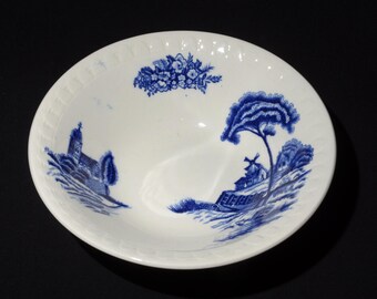 May include: White ceramic bowl with a blue landscape design. The bowl features a rural scene with a church, trees, and a windmill. The rim has a textured, ridged pattern. The bowl is likely for serving food.