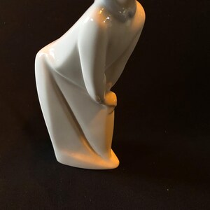 May include: A white porcelain figurine of a person in a long, flowing robe, with their head bowed and hands clasped in front of them.