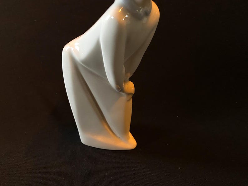 May include: A white porcelain figurine of a person in a long, flowing robe, with their head bowed and hands clasped in front of them.