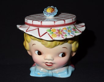 Puede incluir: Un tarro de galletas de cerámica vintage con forma de cabeza de niño que lleva un sombrero de cuadros blancos y rojos con una flor azul en la parte superior. El tarro tiene un diseño pintado de flores y un lazo azul.
