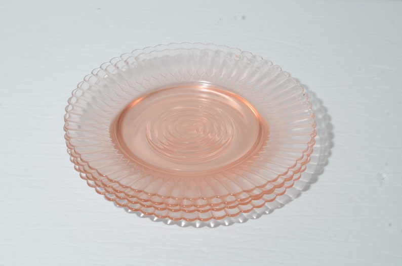 Puede incluir: Una pila de cuatro platos de cristal rosa. Cada plato tiene un borde festoneado y un dise&ntilde;o acanalado que irradia desde el centro. Los platos son de un color rosa suave y transl&uacute;cido, y est&aacute;n apilados.