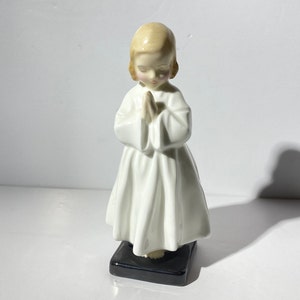May include: A white porcelain figurine of a young girl with blonde hair, wearing a white dress and praying with her hands clasped together. The figurine is standing on a black base.