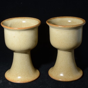 May include: Two beige ceramic goblets with a speckled texture and a brown rim. The goblets have a rounded bowl shape and a pedestal base. The background is black.