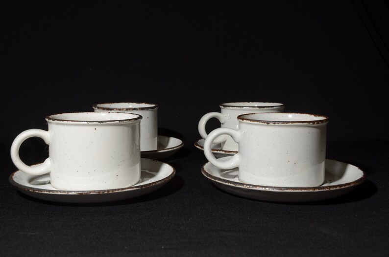 May include: Set of four white ceramic coffee cups with brown rims and speckles. Each cup is paired with a matching saucer.