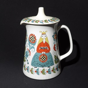 Może przedstawiać: Biały dzbanek ceramiczny z pokrywką, ozdobiony kolorowym wzorem ludowym przedstawiającym kobietę w koronie i czerwono-czarno kratkowanej sukience. Dzbanek ma uchwyt i dzióbek.