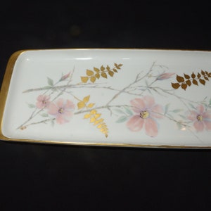 May include: Rectangular white ceramic tray with a gold rim. The tray is decorated with pink flowers, gold leaves, and branches. The floral design is painted on the white surface. The tray is a decorative item.