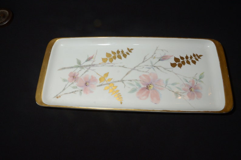 May include: Rectangular white ceramic tray with a gold rim. The tray is decorated with pink flowers, gold leaves, and branches. The floral design is painted on the white surface. The tray is a decorative item.