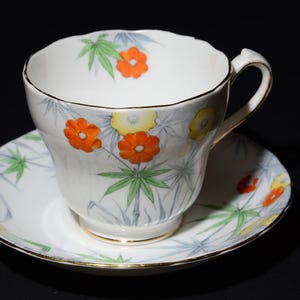 May include: White teacup and saucer set with a floral design. The cup and saucer feature orange and yellow flowers with green leaves and gray bamboo-like stems. The rims are trimmed with gold.