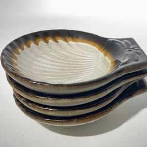 May include: A stack of four ceramic spoon rests. Each rest has a white interior with a textured, fan-like pattern. The rims transition from brown to a dark gray, with a small, decorative floral design on the handle.