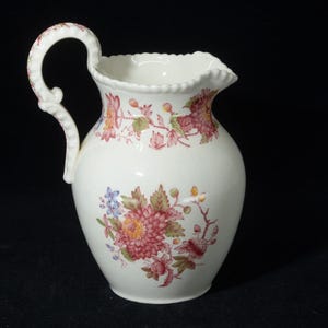 May include: White ceramic pitcher with a floral design in shades of pink, red, blue, and green. The pitcher has a curved handle and a scalloped rim. The floral pattern is repeated around the body of the pitcher.