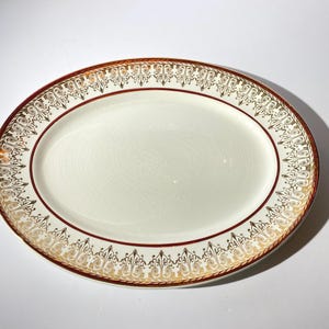 May include: Oval serving platter with a cream-colored center and a decorative border. The border features a gold and burgundy design with intricate patterns. A thin burgundy line separates the center from the border.