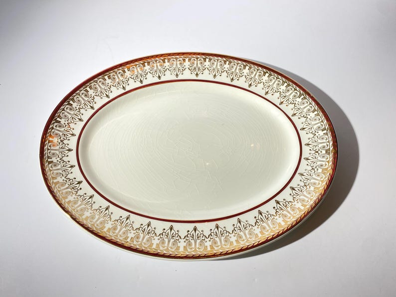 May include: Oval serving platter with a cream-colored center and a decorative border. The border features a gold and burgundy design with intricate patterns. A thin burgundy line separates the center from the border.