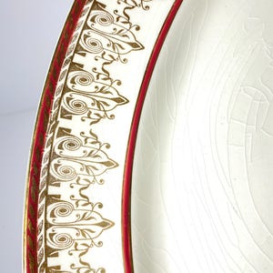 May include: Close-up of a white plate with a decorative gold and red border. The border features an ornate design with gold scrollwork and a red inner band. The plate has a cracked, aged appearance.