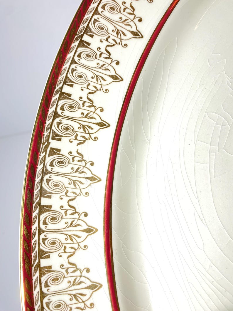 May include: Close-up of a white plate with a decorative gold and red border. The border features an ornate design with gold scrollwork and a red inner band. The plate has a cracked, aged appearance.