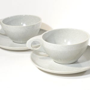 May include: Two grey ceramic teacups and saucers. The cups have a speckled glaze and a rounded shape. The saucers are plain and slightly larger than the cups.
