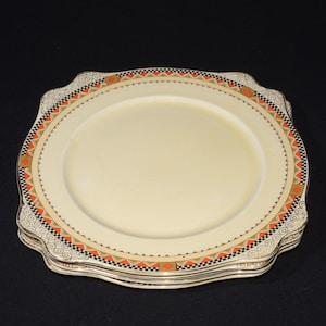 May include: A stack of cream-colored plates with a decorative border featuring orange, black, and gold geometric patterns. The plates have a scalloped edge and are set against a black background.