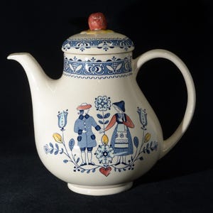 May include: A cream-colored ceramic teapot featuring a blue, red, and yellow folk art illustration of two figures standing among flowers. The lid has a blue patterned border and a small red knob. The teapot has a curved handle and spout.
