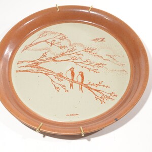 May include: A terracotta-colored ceramic plate with a white center featuring a hand-drawn design of three birds perched on a branch. The design is in orange and the plate has a brown rim.